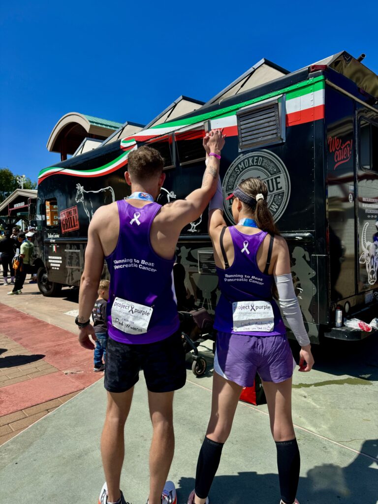 Two Project Purple runners with hands up and clasped together displaying the words "A world without pancreatic cancer" on the back of their singlets