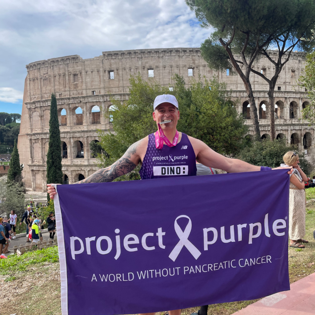 Project Purple Founder Dino Verrelli in a Project Purple singlet, posing in front of the Colosseum. He is holding a Project Purple flag and clenching his Rome Half Marathon finisher medal between his teeth.