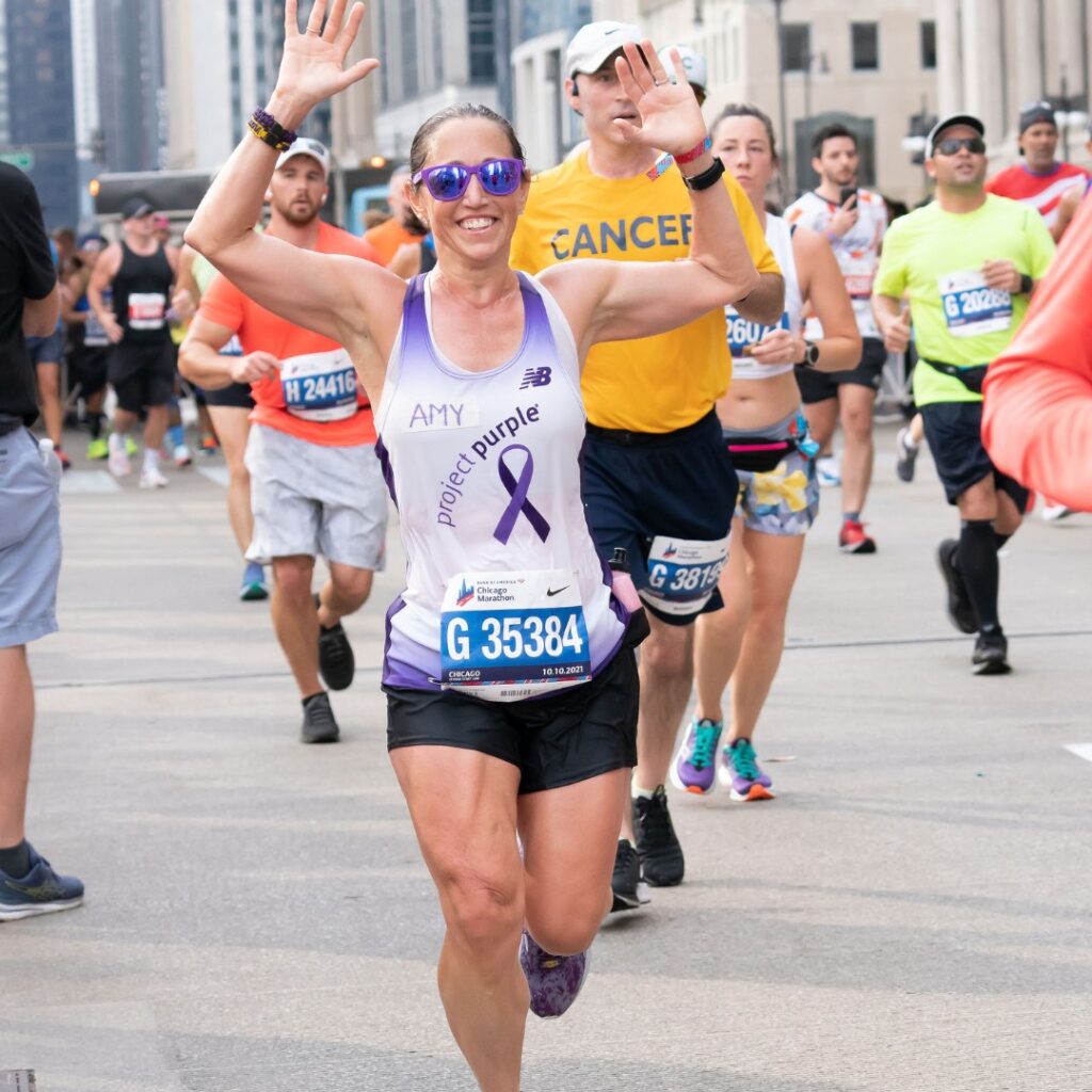 Amy Nugent running and raising her hands in celebration at the 2022 Chicago Marathon where she ran for Project Purple. She is wearing her Project Purple team singlet.